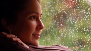 Woman looking out of rainy window with a slight smile on her face.