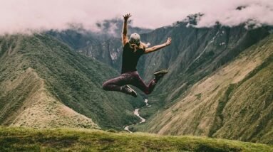 A woman jumping high in the green and lush mountains.