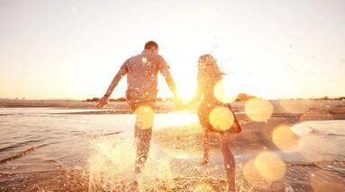 A happy couple running down the edge of the water on a beach during the golden hour.