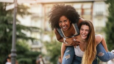 Two happy and confident women having fun in park, one of them riding on the other one's back.