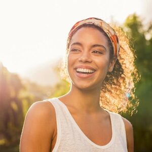 A happy and smiling woman outside with trees and the sun behind her.