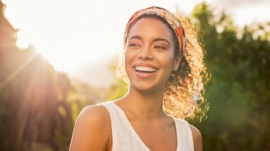 A happy and smiling woman outside with trees and the sun behind her.