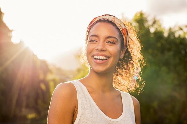 A happy and smiling woman outside with trees and the sun behind her.