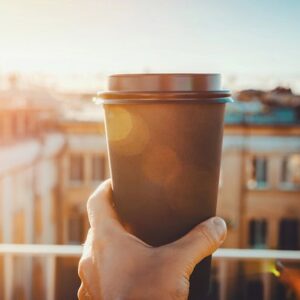 A person on a balcony with hand holding up a cup of coffee towards the sun in in the morning.