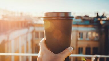 A person on a balcony with hand holding up a cup of coffee towards the sun in in the morning.