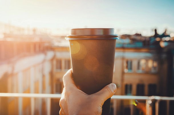 A person on a balcony with hand holding up a cup of coffee towards the sun in in the morning.