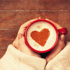 A mug of coffee with a red heart in the foam. The is held by a woman's hands.