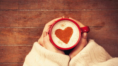 A mug of coffee with a red heart in the foam. The is held by a woman's hands.