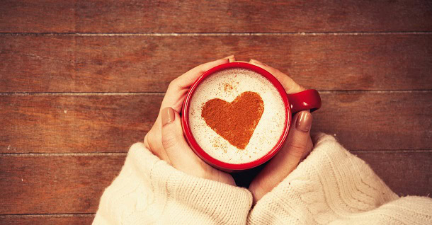 A mug of coffee with a red heart in the foam. The is held by a woman's hands.