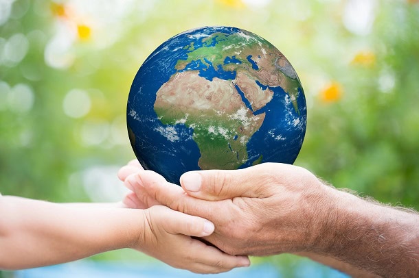 The Earth being held by one young hand and one old hand with a backdrop of green nature.