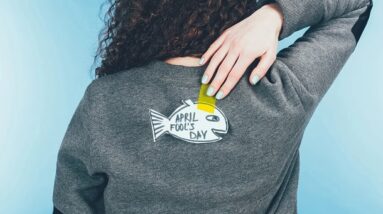 A woman trying to reach a paper sticker in a fish shape on the back on her sweater. The sticker says April Fools Day.