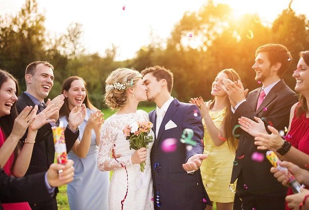 A happy couple kissing on the their wedding day while their friends happily cheer them on.