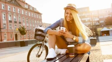 A girl sitting on a bench in a city in summer looking at her phone with a smile.