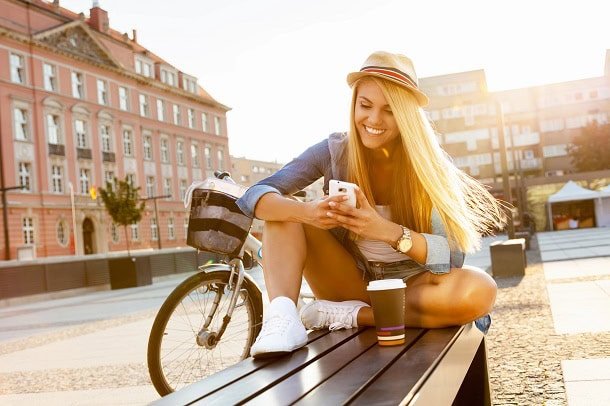 A girl sitting on a bench in a city in summer looking at her phone with a smile.
