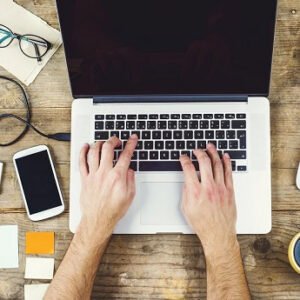 A man working with a laptop with a coffee cup and other things on his desk.