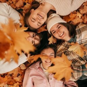 Women lying on autumn leaves and smiling while more leaves fall on them.