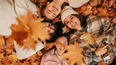 Women lying on autumn leaves and smiling while more leaves fall on them.