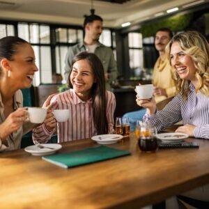 Three women laughing while sitting in a cafe with coffee cups in their hands.
