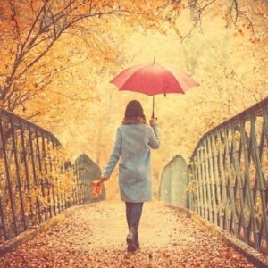 A woman with an umbrella walking over a bridge with fall colors all around her.