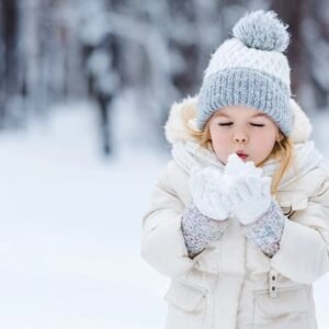 A small girl blowing at snow in her hands while standing in a forest in winter.