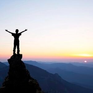 A man at a top of a mountain stretching his hands upward in celebration during sunset.