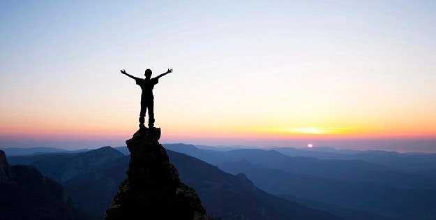 A man at a top of a mountain stretching his hands upward in celebration during sunset.