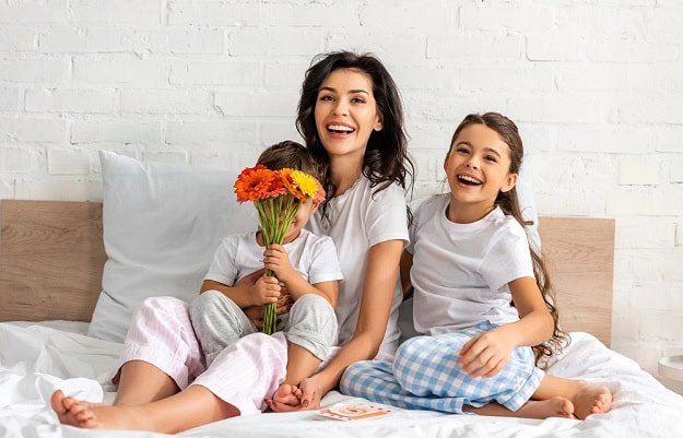 A happy mother with her two smiling children in bed and a bouquet of flowers.